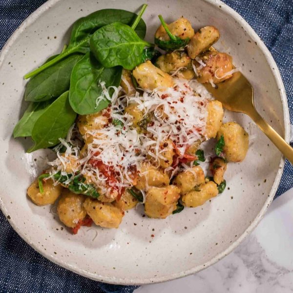 A white ceramic bowl sitting on a blue napkin. In the bowl is a serving of vegetarian baked gnocchi with extra spinach leaves and a gold fork sitting in the dish.