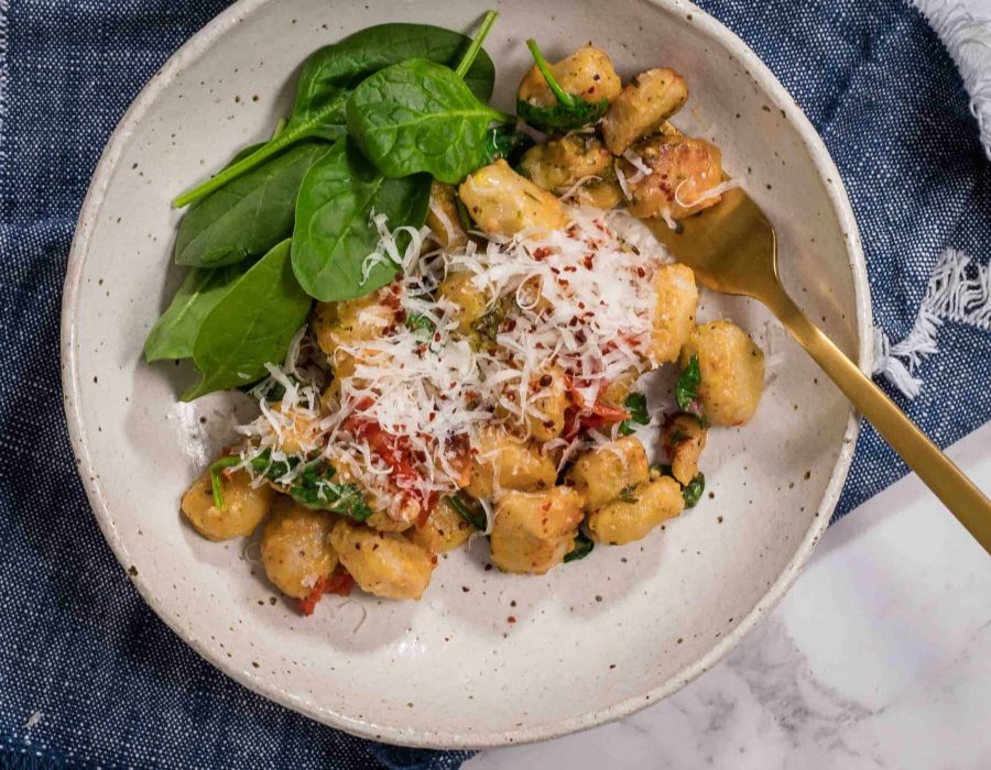 A white ceramic bowl sitting on a blue napkin. In the bowl is a serving of vegetarian baked gnocchi with extra spinach leaves and a gold fork sitting in the dish.