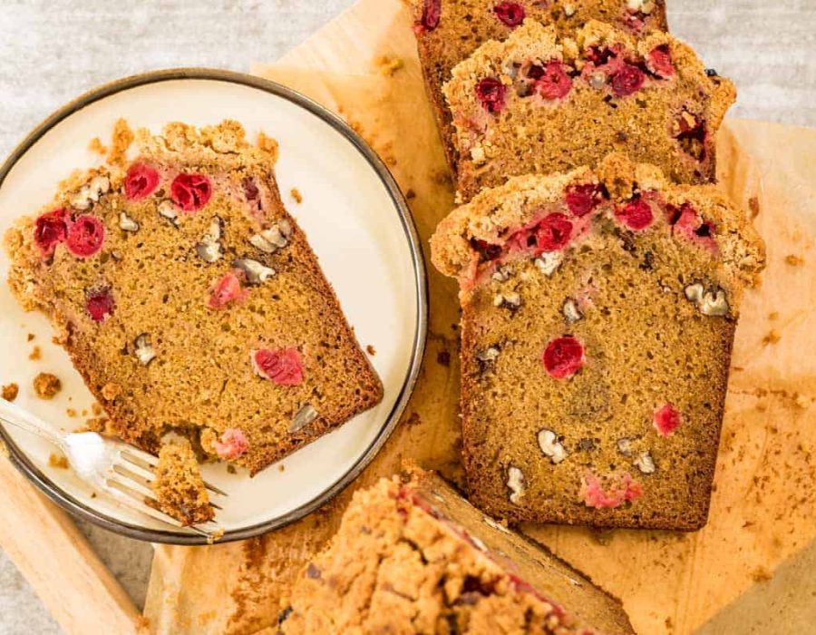 A cream colored plate with a slice of orange cranberry cake and a silver fork. There are slices of the same cake alongside the plate on a wooden board.