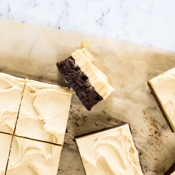 Square-cut dark chocolate brownies topped with a thick swirl of light-tan frosting rest on brown parchment paper for chocolate and peanut butter brownies.