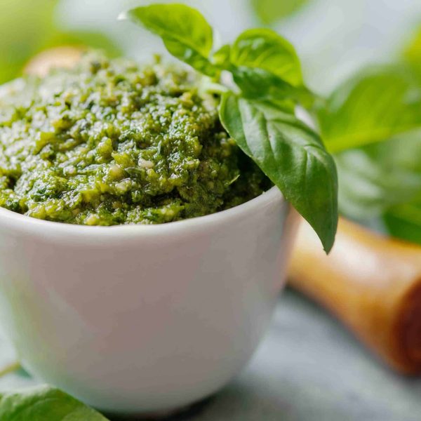 Freshly made pistachio pesto with basil in a small white bowl, with a wooden spoon and more fresh basil in the background.