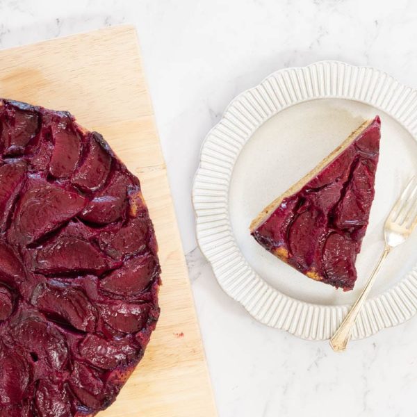 Slice of golden-brown upside down cake topped with deep purple cooked plums is served on a white ridged plate with a silver fork.