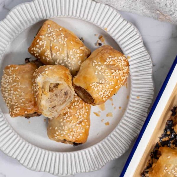 A close-up, overhead shot of five golden-brown pork and apple pastry rolls piled onto a ribbed white ceramic plate next to a blue-rimmed baking pan, with one roll cut in half to show the cooked meat and apple filling inside for sausage rolls.