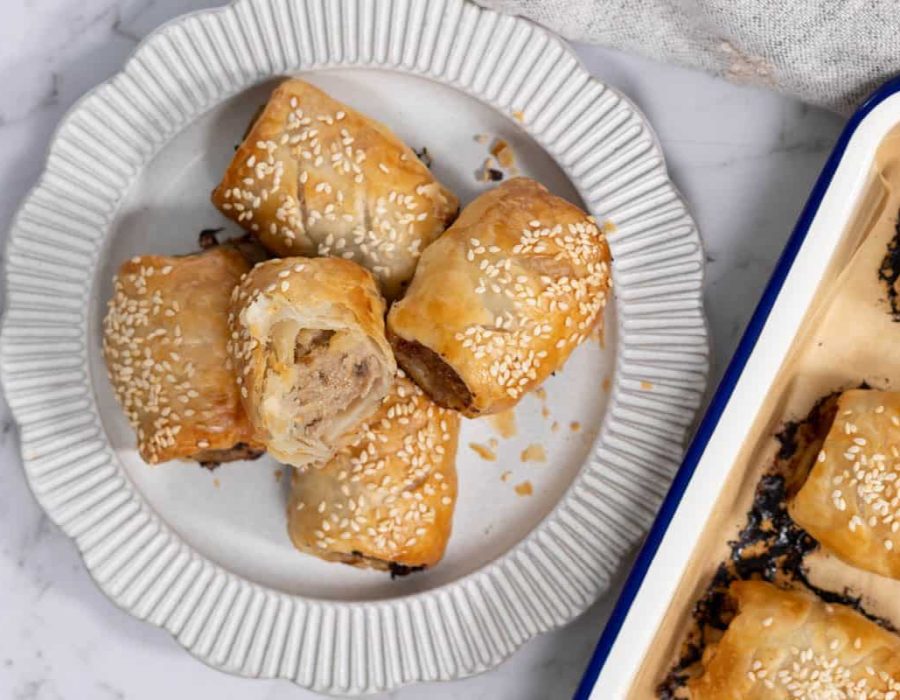 A close-up, overhead shot of five golden-brown pork and apple pastry rolls piled onto a ribbed white ceramic plate next to a blue-rimmed baking pan, with one roll cut in half to show the cooked meat and apple filling inside for sausage rolls.