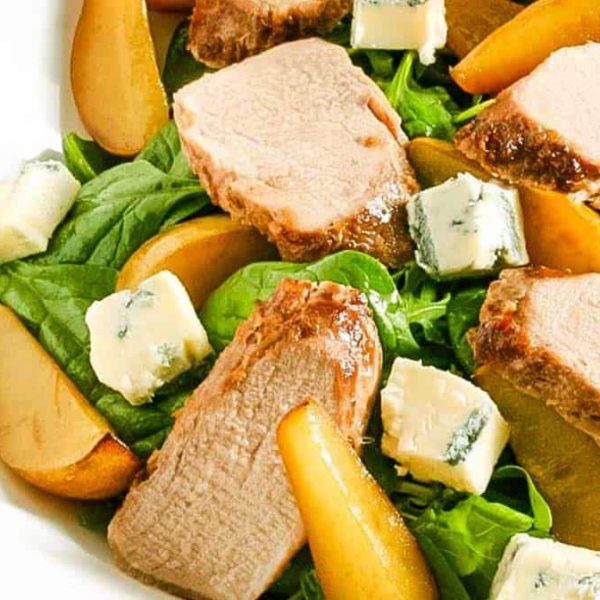 A white bowl with a salad of roasted pork and slices of pear with salad greens and gorgonzola cheese chunks
