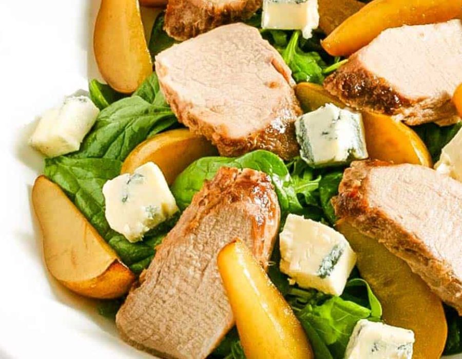 A white bowl with a salad of roasted pork and slices of pear with salad greens and gorgonzola cheese chunks
