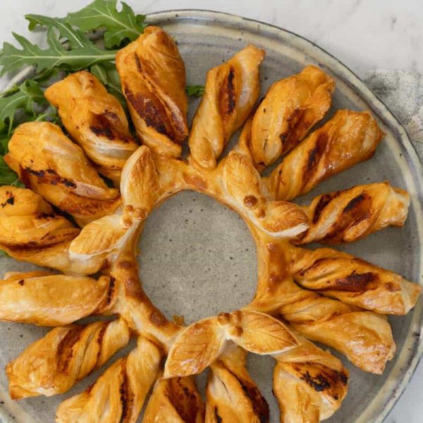 A red pesto and puff pastry wreath, made by twisting strips of pastry attached to a central ring, then baking. The wreath is served on a gray ceramic plate with salad leaves for garnish.