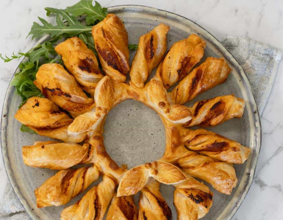 A red pesto and puff pastry wreath, made by twisting strips of pastry attached to a central ring, then baking. The wreath is served on a gray ceramic plate with salad leaves for garnish.