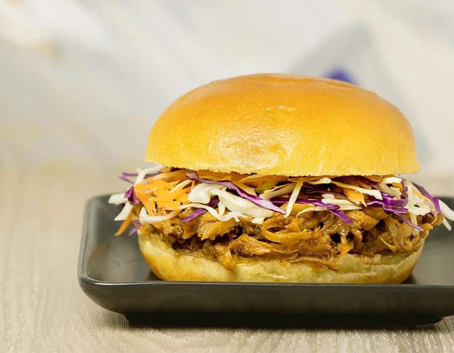 A burger bun with pulled pork and coleslaw on a curved black plate on a light wood counter