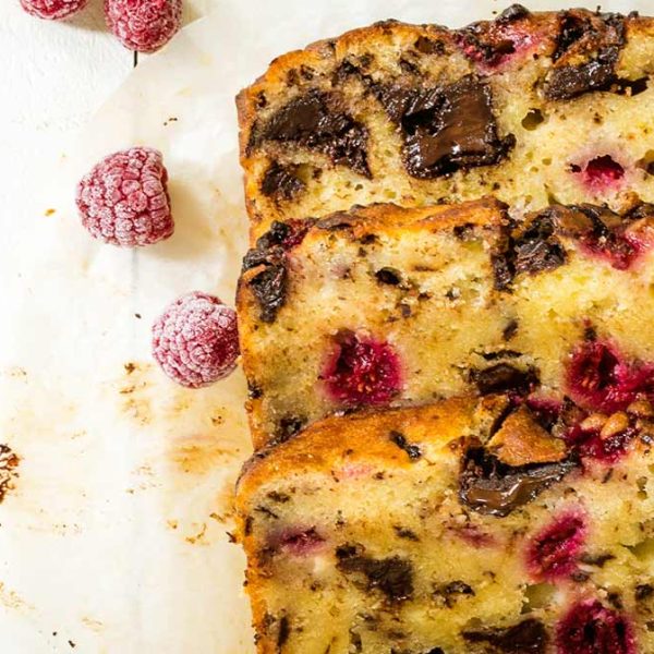 Three slices of raspberry chocolate loaf cake, lying at an angle on parchment paper with a few frozen raspberries scattered around them.