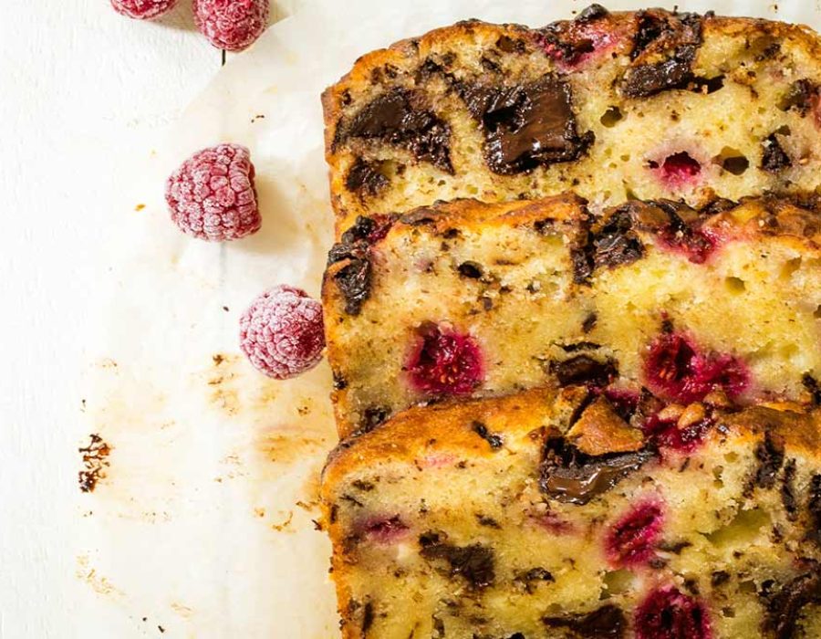 Three slices of raspberry chocolate loaf cake, lying at an angle on parchment paper with a few frozen raspberries scattered around them.