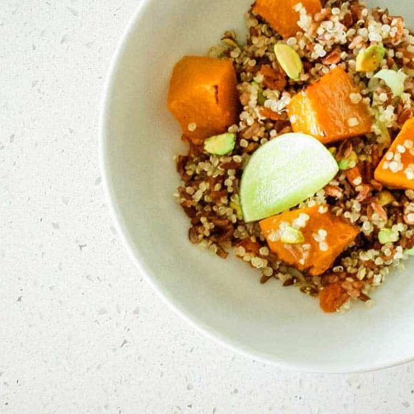 A close up image of a deep white bowl red rice, quinoa, pumpkin chunks, nuts and a wedge of lime, on a speckled counter top