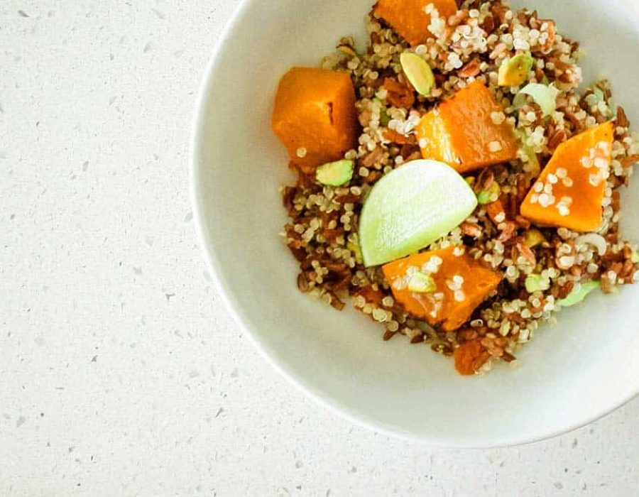 A close up image of a deep white bowl red rice, quinoa, pumpkin chunks, nuts and a wedge of lime, on a speckled counter top