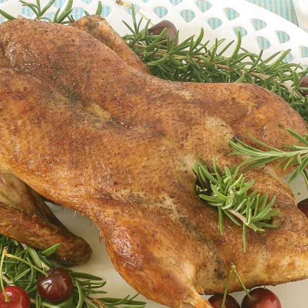 A white platter with cutout edges on a striped cloth with a whole roast duck, rosemary sprigs and cherries
