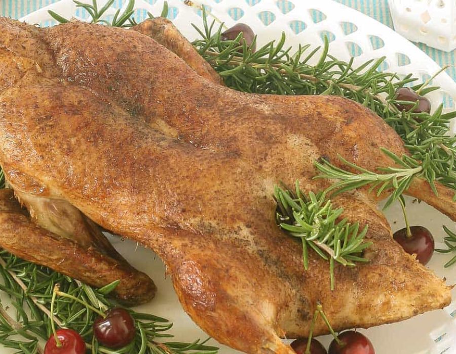 A white platter with cutout edges on a striped cloth with a whole roast duck, rosemary sprigs and cherries