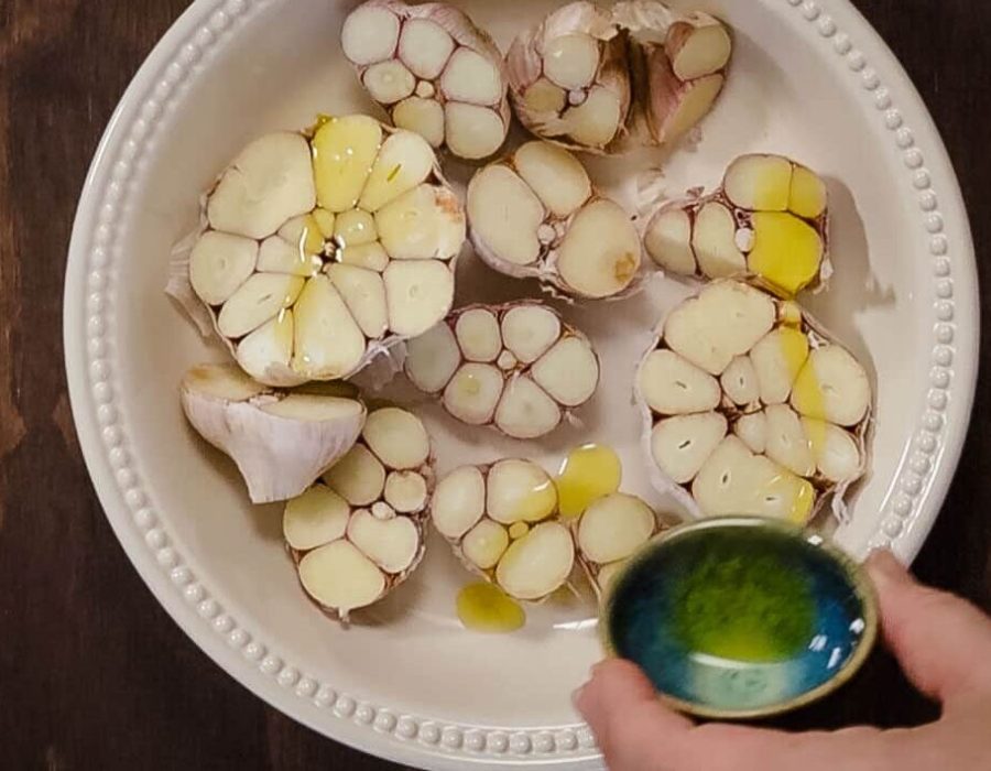a round ceramic baking dish holds several heads of garlic which have been halved; a hand holds a small dish of olive oil which is being drizzled over the garlic.