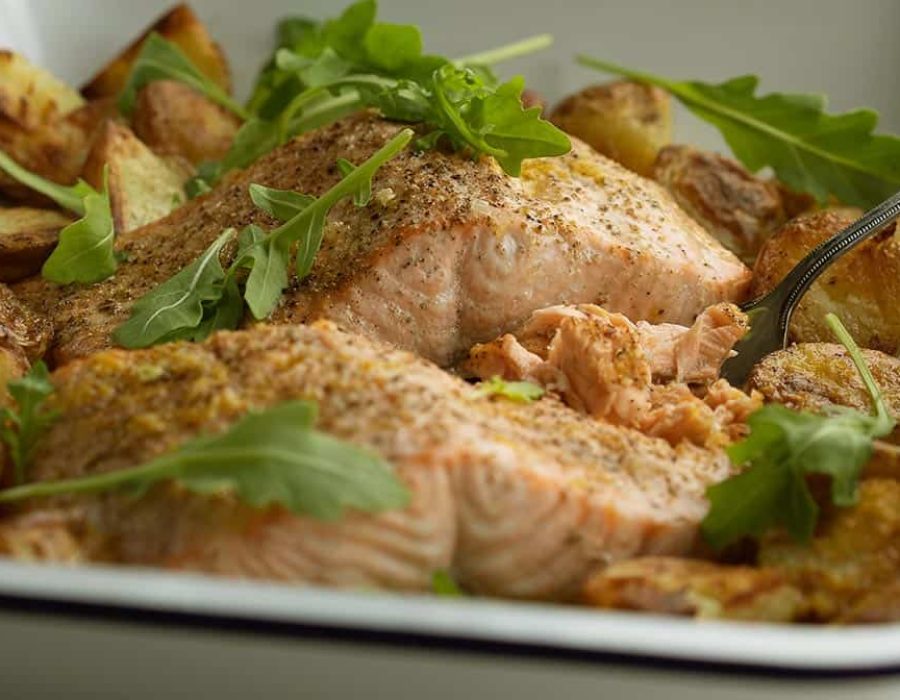 A close up view of the texture of roasted salmon with lemon and burnt butter in a baking dish along with rocket and roast potatoes