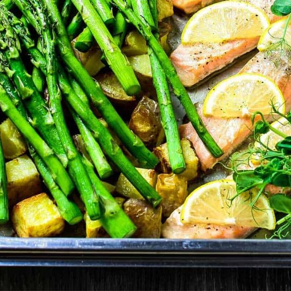 Steam oven salmon in a stainless steel roasting dish with slices of lemon, asparagus, roast potatoes and pea shoots.