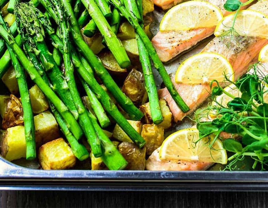 Steam oven salmon in a stainless steel roasting dish with slices of lemon, asparagus, roast potatoes and pea shoots.