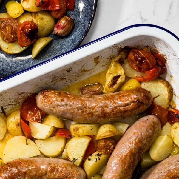Overhead landscape view of a white baking dish containing roasted sausages, golden potatoes, and blistered tomatoes next to a dark blue plate with a served portion for a sausage bake.
