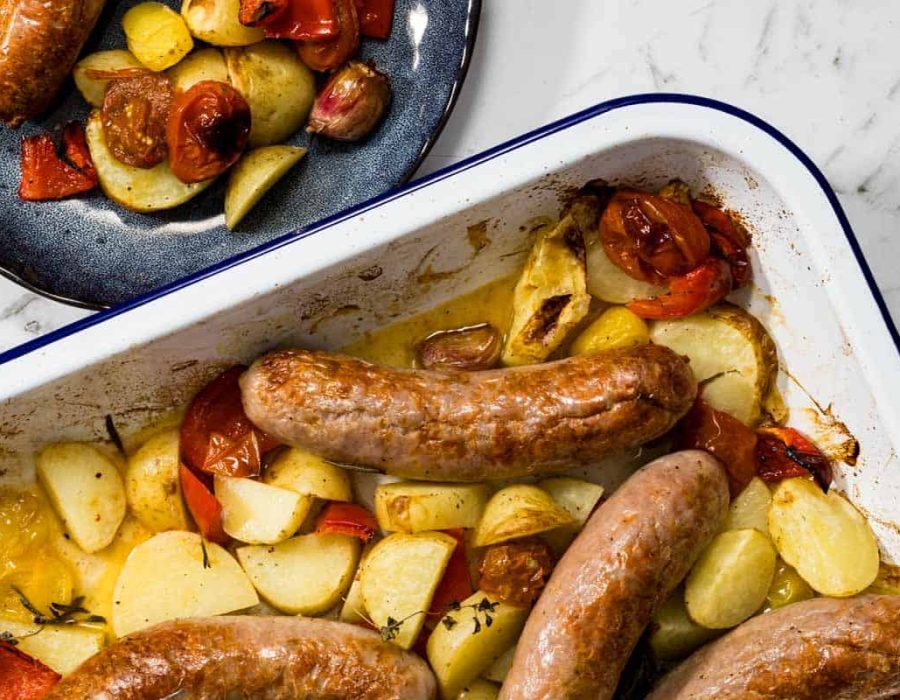 Overhead landscape view of a white baking dish containing roasted sausages, golden potatoes, and blistered tomatoes next to a dark blue plate with a served portion for a sausage bake.