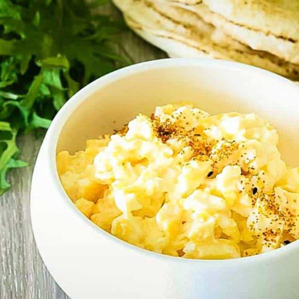 A small white bowl with fluffy, seasoned scrambled eggs with tortillas and rocket in the background.