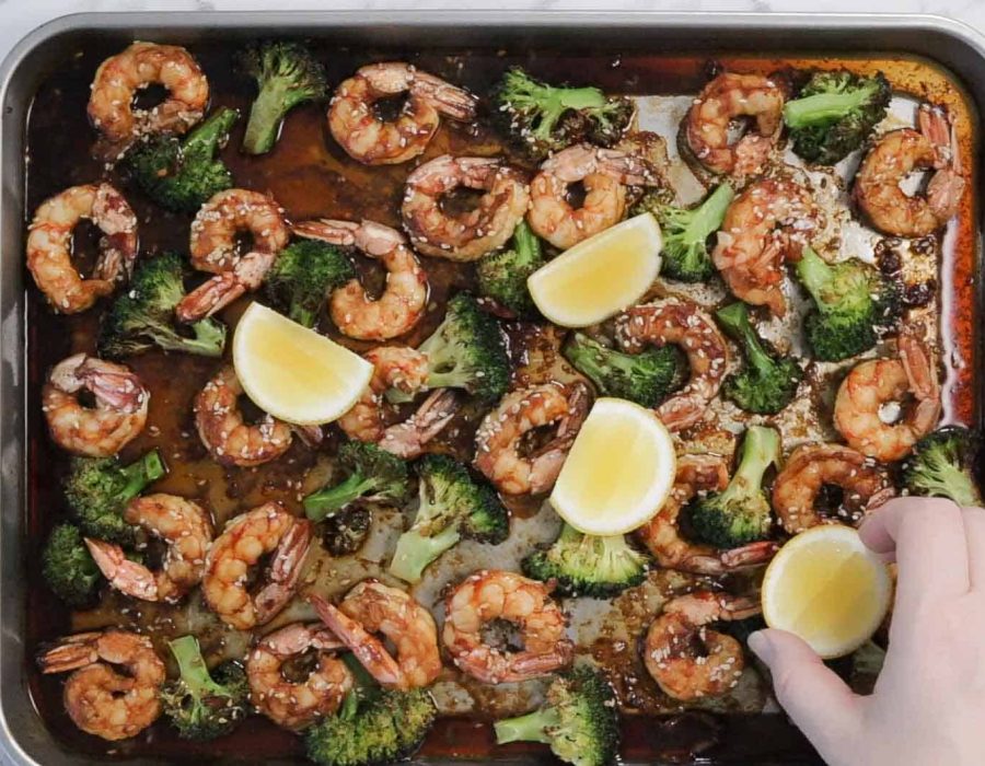 A sheet pan full of roasted broccoli and shrimp in soy ginger sauce. A hand places fresh lemon wedges around the pan for serving.