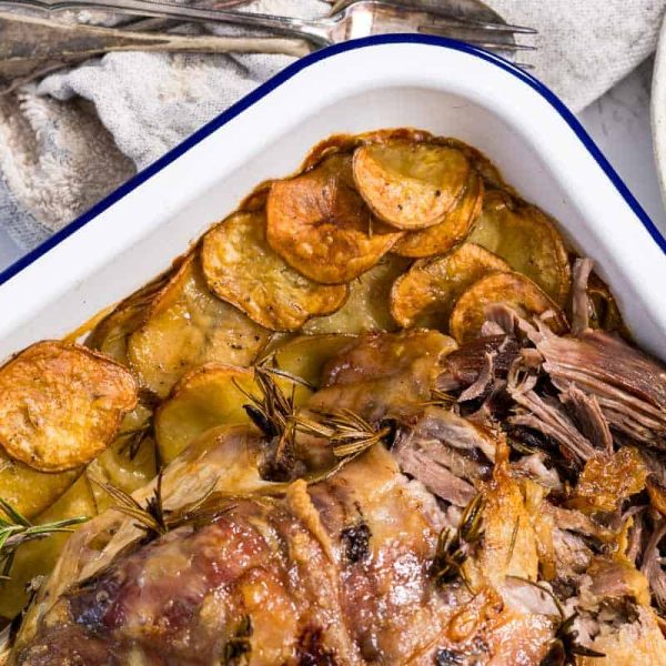 A close-up, angled view of roasted lamb leg with tender meat falling away from the bone, surrounded by golden-brown crispy potato slices in a white enamel pan.