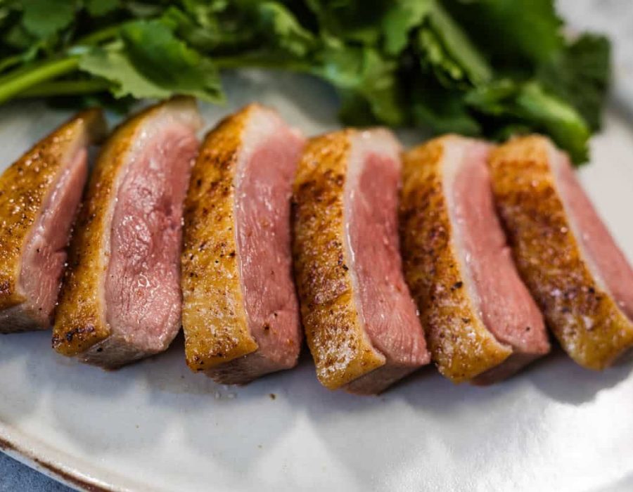 A close-up image of sliced crispy skinned sous vide duck breast, served on a white plate with cilantro leaves in the background.