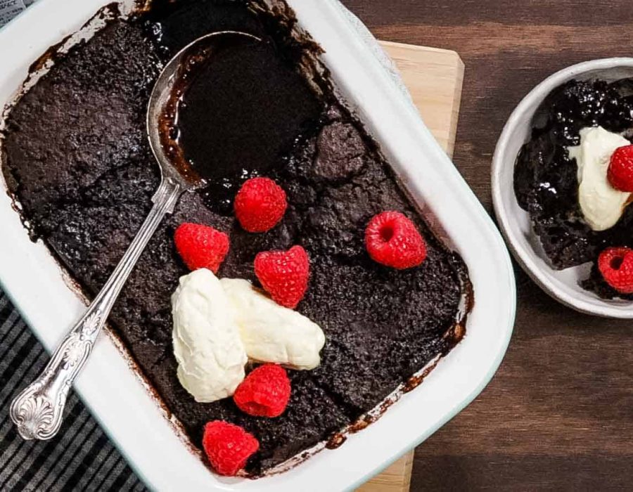 A rectangular, white enamel baking dish is filled with freshly baked steam oven chocolate pudding and garnished with thick cream and fresh raspberries. One serve has been scooped out of the dish and is in a round white bowl alongside.