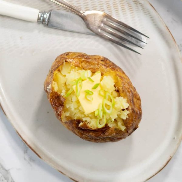 A single steam baked potato is split open on a light ceramic plate, revealing a fluffy yellow interior topped with a square melting pat of butter and sliced green onions next to a silver fork and knife.