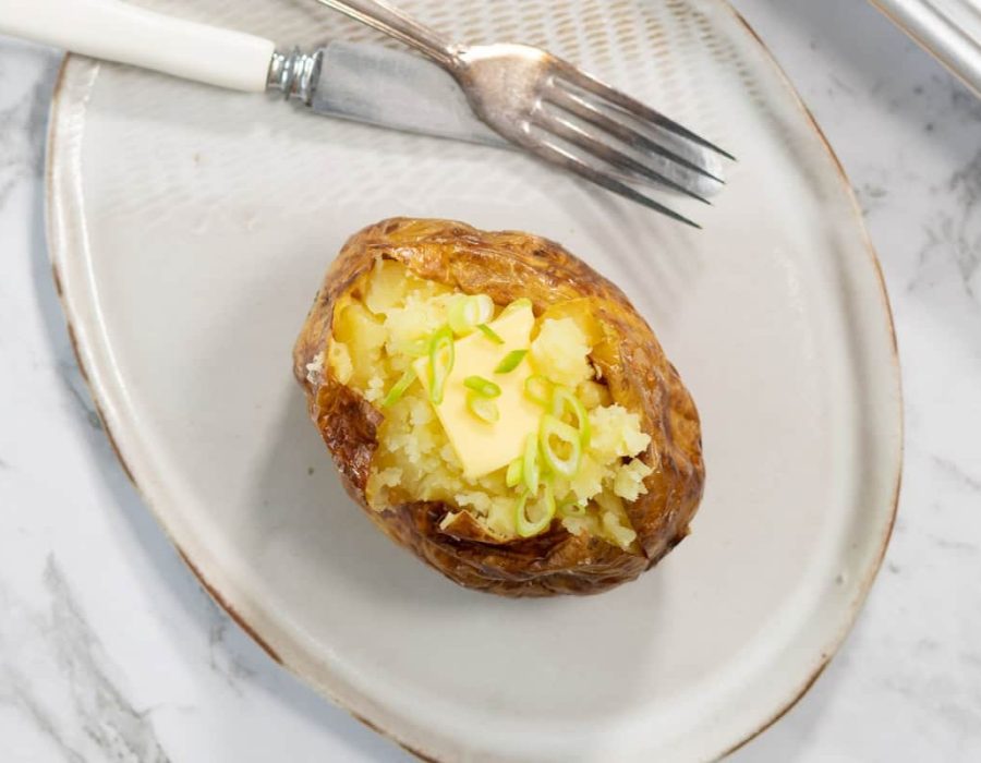 A single steam baked potato is split open on a light ceramic plate, revealing a fluffy yellow interior topped with a square melting pat of butter and sliced green onions next to a silver fork and knife.