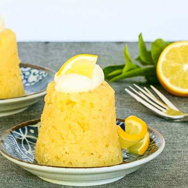 two individual steamed lemon puddings garnished with cream and lemon with dessert fork and a halved lemon on a dark surface