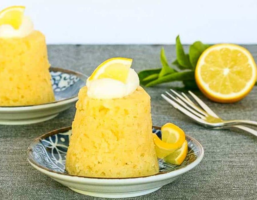 two individual steamed lemon puddings garnished with cream and lemon with dessert fork and a halved lemon on a dark surface