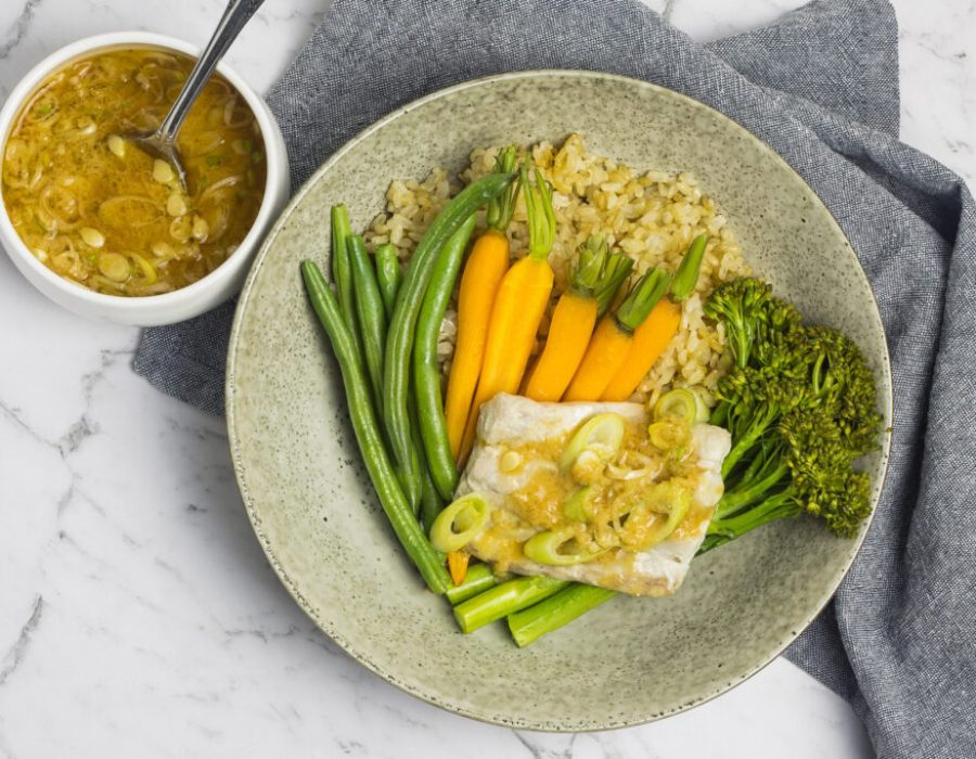 A zoomed out picture of a grey speckled plate with steamed fish, mixed vegetables with a bowl of miso dressing