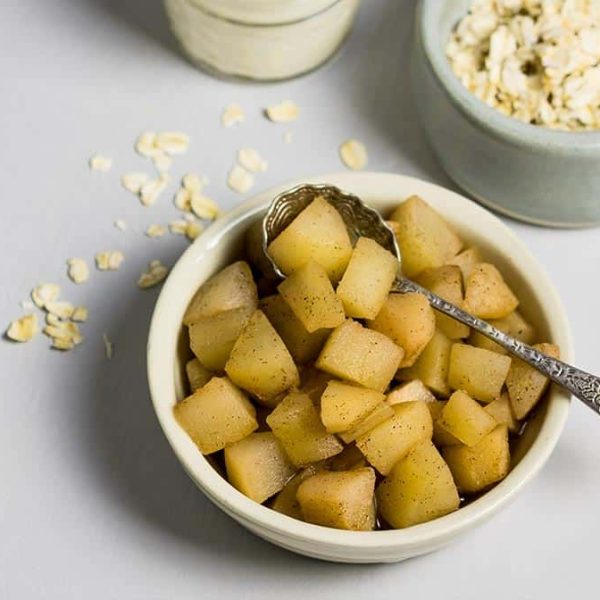 a small white bowl with a decorative spoon, filled with stewed diced fruit with a ramekin of rolled oats and a jar of yoghurt on the counter