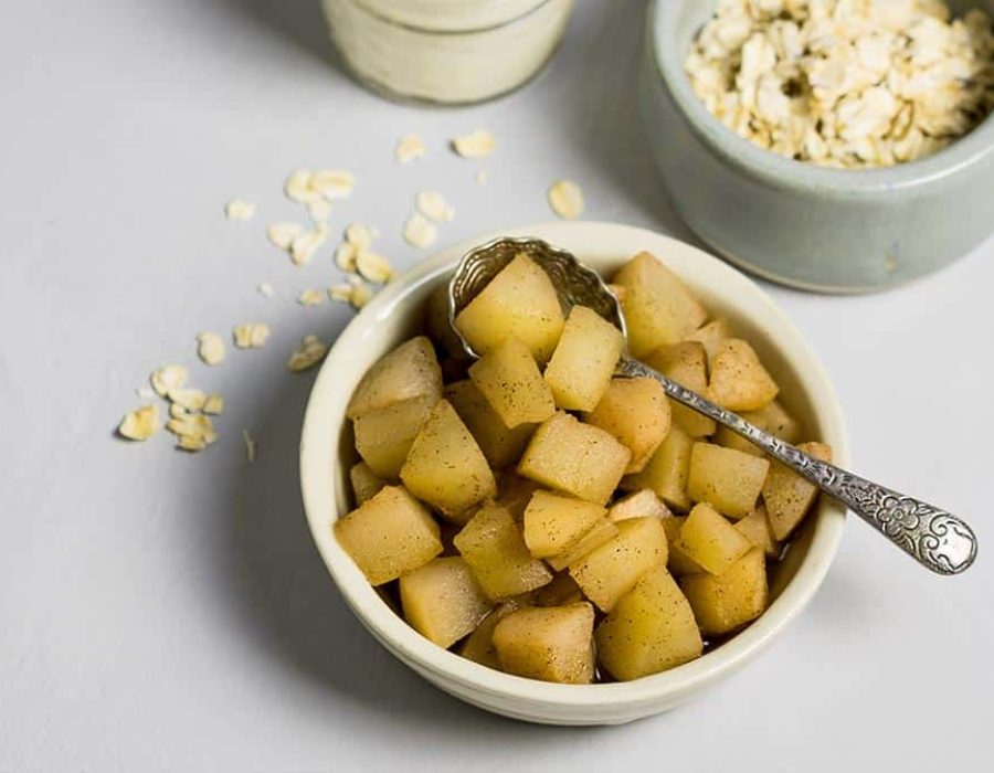 a small white bowl with a decorative spoon, filled with stewed diced fruit with a ramekin of rolled oats and a jar of yoghurt on the counter