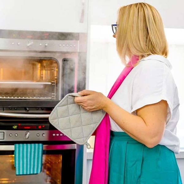 A woman with blonde hair, wearing a white shirt and green skirt, stands in front of an open steam oven. She holds a pot mitt, ready to remove a hot pan.