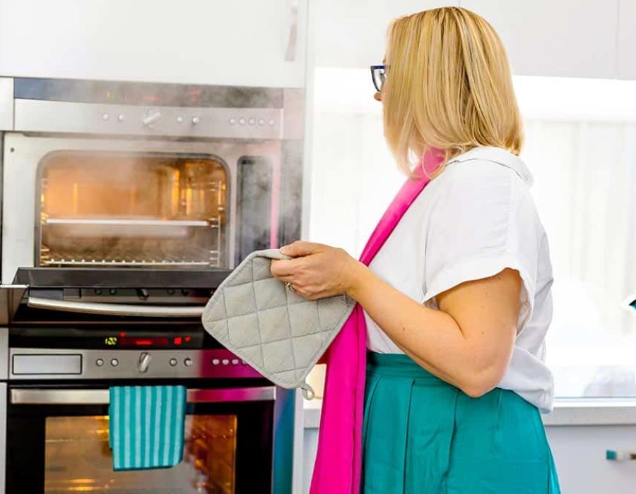A woman with blonde hair, wearing a white shirt and green skirt, stands in front of an open steam oven. She holds a pot mitt, ready to remove a hot pan.