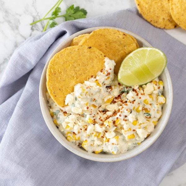 creamy corn appetizer topped with crumbled cheese, chili seasoning, and a fresh lime wedge, served with round tortilla chips in a ceramic bowl over a blue linen napkin
