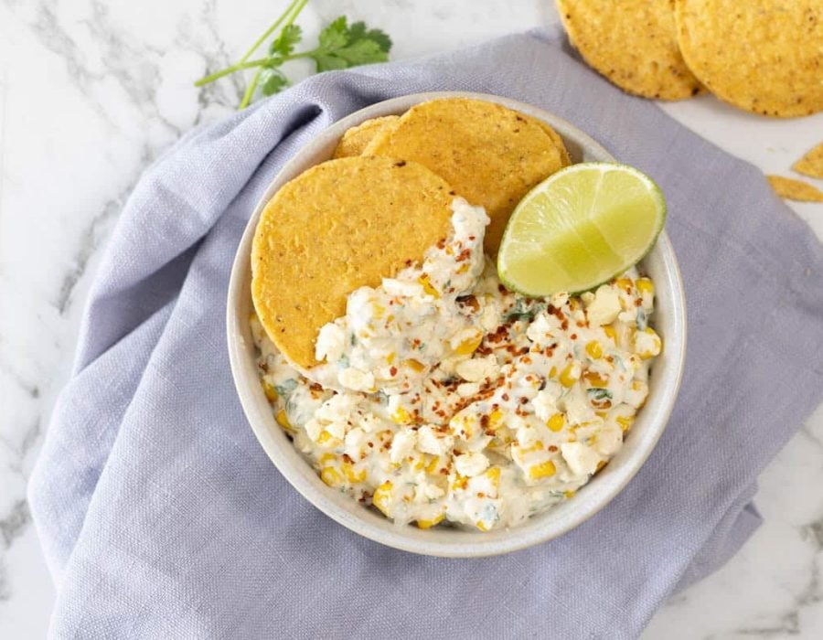 creamy corn appetizer topped with crumbled cheese, chili seasoning, and a fresh lime wedge, served with round tortilla chips in a ceramic bowl over a blue linen napkin
