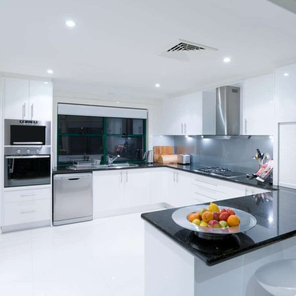 A modern, glossy white kitchen with black countertops. The kitchen is a U shape with built in steam oven on the far side and a peninsula with food on it in the foreground.