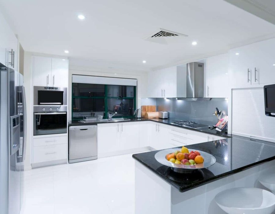 A modern, glossy white kitchen with black countertops. The kitchen is a U shape with built in steam oven on the far side and a peninsula with food on it in the foreground.